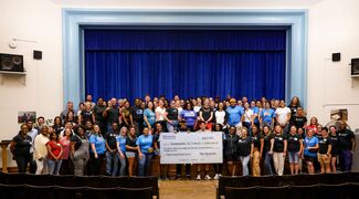 Large group of students gathered in an auditorium smiling and holding an oversized check with the donation
